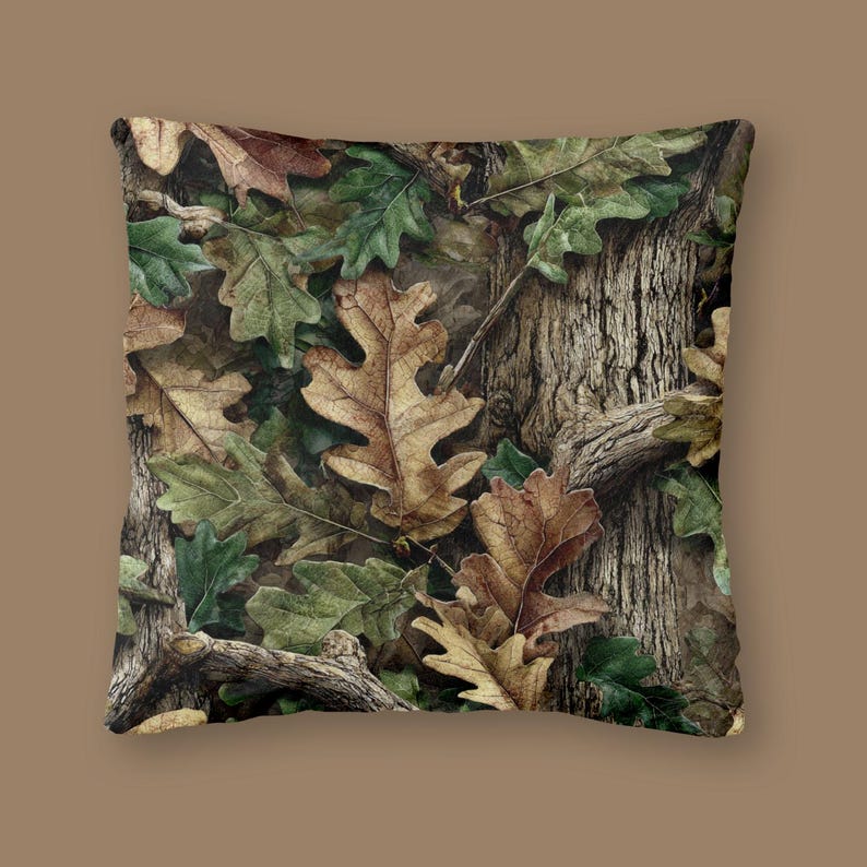 May include: Square cushion with a camouflage design of brown and tan oak leaves, green leaves, and tree bark. The pillow has a neutral background and is suitable for home decor.