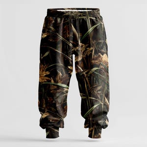 May include: Camouflage-patterned sweatpants featuring a blend of brown, green, and tan hues. The design incorporates leaves and tall grass, creating a woodland camouflage effect. The pants have an elastic waistband and cuffs.