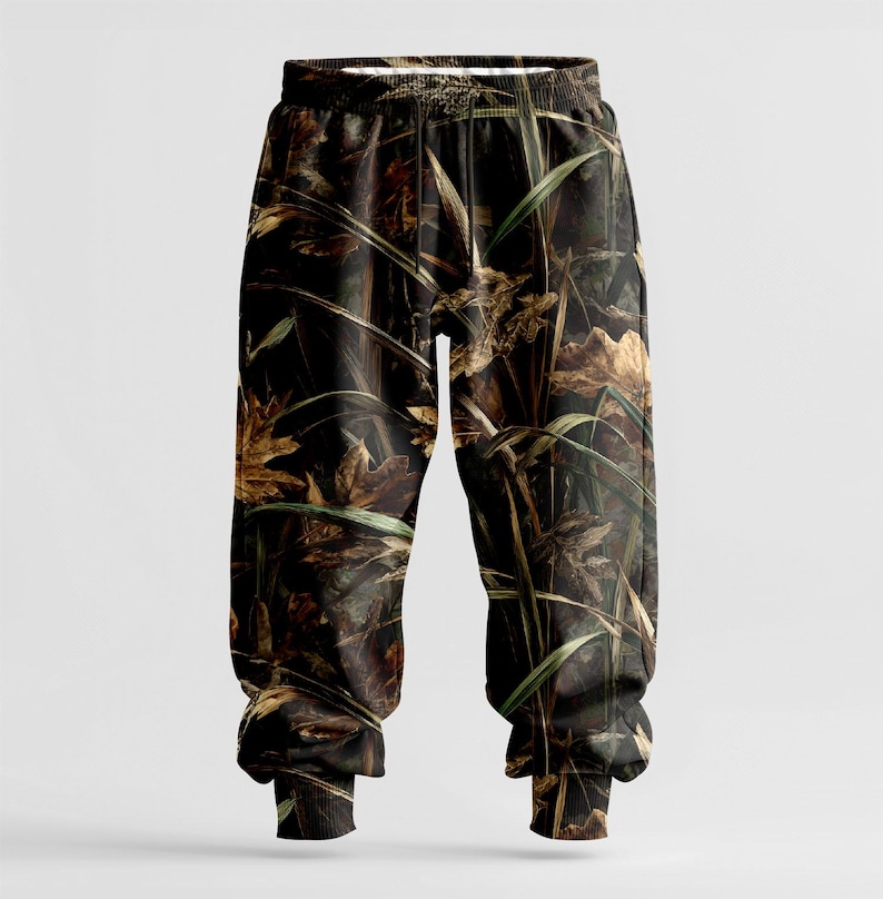 May include: Camouflage-patterned sweatpants featuring a blend of brown, green, and tan hues. The design incorporates leaves and tall grass, creating a woodland camouflage effect. The pants have an elastic waistband and cuffs.