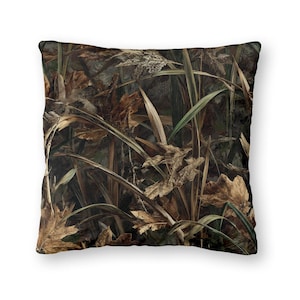 May include: Square throw pillow with a camouflage pattern. The design features a mix of brown, green, and tan colors, mimicking a natural outdoor environment with leaves and foliage. The pillow is likely for home decor.