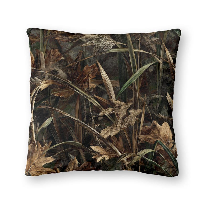 May include: Square throw pillow with a camouflage pattern. The design features a mix of brown, green, and tan colors, mimicking a natural outdoor environment with leaves and foliage. The pillow is likely for home decor.