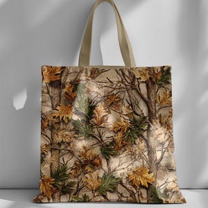 K&ouml;nnte beinhalten: Beige Tragetasche mit einem Tarnmuster aus &Auml;sten und Bl&auml;ttern in Braun-, Gr&uuml;n- und Gelbt&ouml;nen. Die Tasche hat einen hellbeigen Griff.