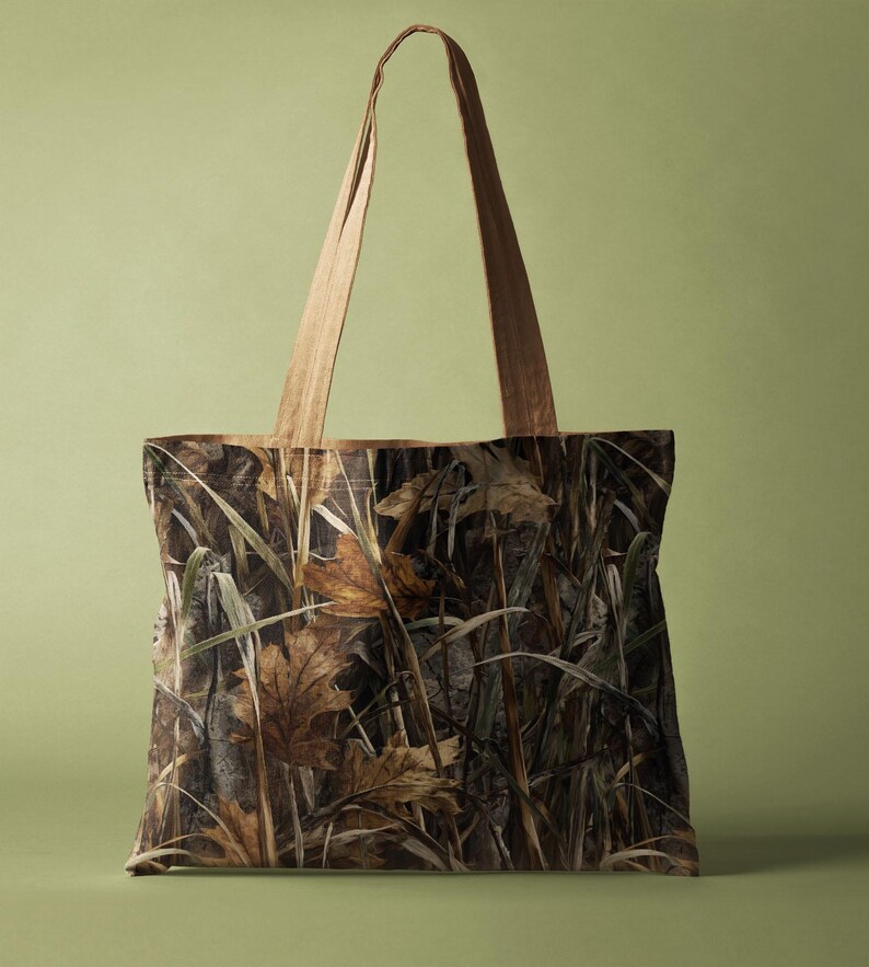 May include: A tote bag featuring a camouflage pattern of brown and tan leaves and grasses. The bag has light brown handles and a rectangular shape. The background is a solid light green.