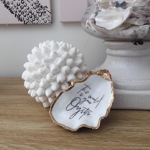 May include: White ceramic oyster shell dish with gold trim and the text "The world is your oyster" written in black calligraphy.