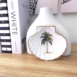 May include: White seashell-shaped dish with a gold rim and a painted palm tree design. The dish is in front of a white vase. Books with the word "WHITE" are visible in the background.