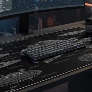 Op de afbeelding: Een zwarte bureaumat met witte schema's van verschillende ruimtevaartuigen, een zwart toetsenbord en een computermuis. Een dampende zwarte mok en een klein beeldje staan ook op het bureau. Het bureau is van hout.