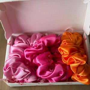 Puede incluir: Una caja de coleteros de satén en tonos rosa y naranja. Los coleteros están colocados dentro de una caja blanca con un estampado de lazo rosa. Los coleteros están hechos de una tela brillante y suave.
