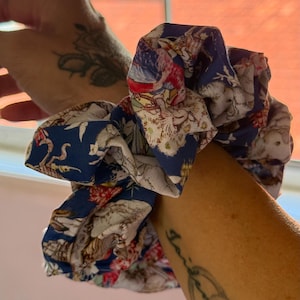 May include: A large, fabric scrunchie with a navy blue background and a detailed floral and animal print in shades of red, white, and beige. The scrunchie is gathered and voluminous, suitable for various hairstyles.