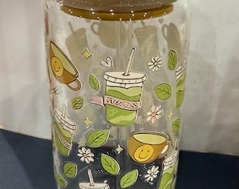 Puede incluir: Un vaso de vidrio transparente con tapa de bambú y pajita transparente. El vaso está decorado con ilustraciones de bebidas matcha verdes, tazas de aguacate, hojas y flores. El diseño se repite alrededor del vaso.