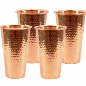 May include: Four copper-colored drinking glasses with a hammered texture. The glasses have a wide rim and a slightly tapered shape. The copper color gives the glasses a warm, inviting appearance. These glasses are suitable for water, juice, or other beverages.