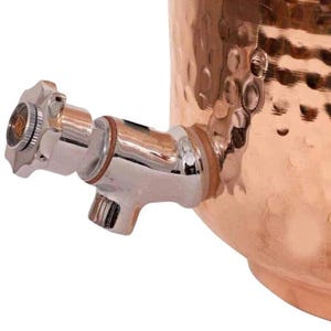 May include: Close-up of a copper beverage dispenser with a polished chrome spigot. The dispenser has a hammered texture and a shiny, metallic finish. The spigot features a star-shaped handle.