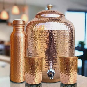 Puede incluir: Un juego de dispensador de agua de cobre con textura martillada. El juego incluye un recipiente grande para agua con tapa, una botella de agua y dos vasos. El dispensador tiene un grifo para verter fácilmente.