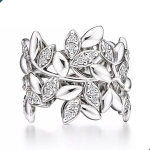 Pode incluir: Um anel de prata com um design de folhas. O anel é adornado com pequenas pedras preciosas brilhantes. As folhas são dispostas em um padrão contínuo ao redor da banda, criando uma estética inspirada na natureza. O anel está em um fundo branco.