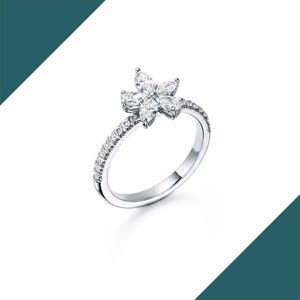 May include: A silver ring featuring a flower design. The flower is composed of six marquise-cut gemstones. The band is encrusted with small, round gemstones. The ring is set against a white background.