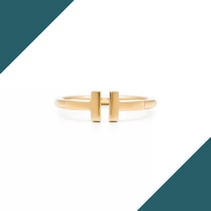 May include: A gold-colored ring with a minimalist design. The ring features a horizontal bar with two vertical bars, forming a stylized 'T' shape. The band is smooth and polished, reflecting light. The ring is a piece of jewelry.