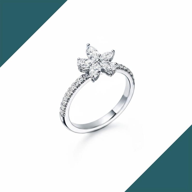 May include: A silver ring featuring a flower design. The flower is composed of six marquise-cut gemstones. The band is adorned with small, round gemstones. The ring is set against a white background.