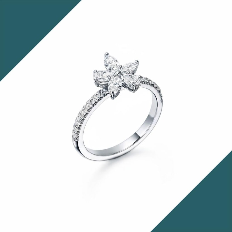 May include: A silver ring featuring a flower design. The flower is composed of six marquise-cut gemstones. The band is encrusted with small, round gemstones. The ring is set against a white background.