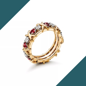 Op de afbeelding: Een goudkleurige ring met een herhalend patroon van rode en witte edelstenen. De ring heeft een uniek ontwerp met een X-vormig patroon tussen de edelstenen. Het is een sieraad.