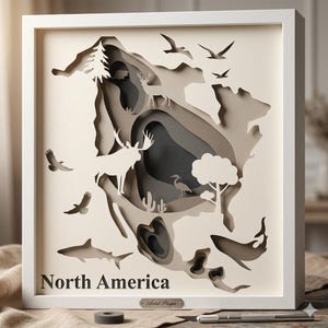 May include: A framed, layered paper art piece depicting North America. The artwork features cut-out silhouettes of wildlife, including a moose, deer, birds, and a whale. The text "North America" is displayed at the bottom, with a small artist plaque.