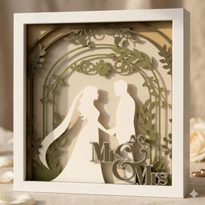 May include: A framed wedding shadow box featuring a bride and groom silhouette under a floral arch. The words "Mr & Mrs" are displayed in silver. The frame is white, and the background has a layered paper design with green and beige tones.