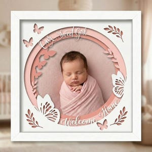 Peut inclure: Un cadre carré blanc contenant une découpe en papier superposée avec un centre circulaire affichant une photo de nourrisson emmailloté. La découpe présente des papillons et des feuilles roses et blancs avec le texte Our Sweet Girl Welcome Home.