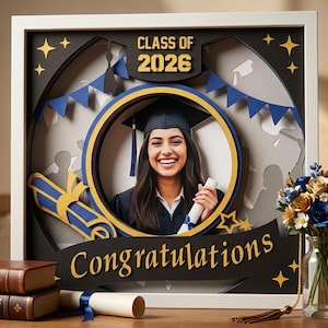 May include: A framed graduation photo featuring a smiling graduate in a cap and gown, holding a diploma. The frame includes the text "Class of 2026" and "Congratulations" with blue and gold accents. A bouquet of flowers and books are in the foreground.