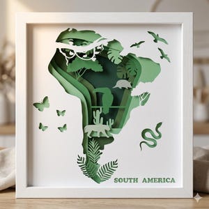May include: A framed paper cut art piece depicting the continent of South America. The artwork features layers of green paper, creating a 3D effect with images of animals, plants, and the text "SOUTH AMERICA" at the bottom.