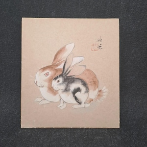 Könnte beinhalten: Ein Aquarellgemälde von zwei Kaninchen. Ein Kaninchen ist braun und weiß, das andere ist schwarz und weiß. Die Kaninchen sitzen eng beieinander auf einem beigefarbenen Hintergrund. Kleine rote und schwarze japanische Schriftzeichen befinden sich in der oberen rechten Ecke.