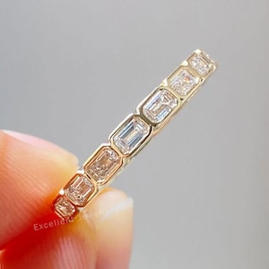 Puede incluir: Un anillo de banda de oro con una serie de piedras preciosas de corte rectangular. El anillo se sostiene entre los dedos sobre un fondo gris suave. Las piedras preciosas son claras y reflejan la luz, y la banda tiene un acabado pulido.