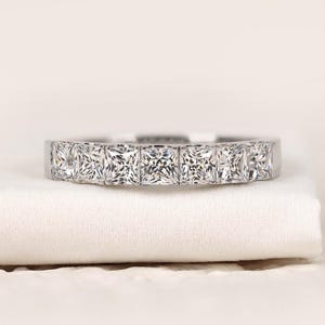 May include: A silver band ring with a line of square-cut, clear gemstones. The ring is displayed on a white surface, highlighting its polished metal and sparkling stones. The design is simple and elegant, suitable for various occasions.