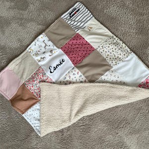 Può includere: Una coperta patchwork per bambini con una varietà di motivi e colori di tessuto, tra cui quadrati floreali, a righe e a tinta unita. La coperta presenta il nome "Esmée" ricamato in nero. Il lato inferiore è in morbido materiale sherpa color crema.