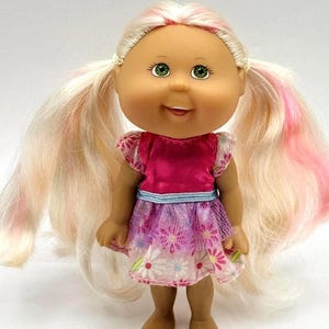 Könnte beinhalten: Eine Vintage-Puppe mit blonden Zöpfen mit rosa Strähnen. Die Puppe hat grüne Augen und trägt ein rosa Oberteil und einen Blumenrock mit blauem Bund. Die Puppe steht auf einer weißen Oberfläche.