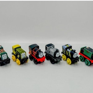 May include: Six miniature toy train engines in a row, each with a different colour scheme and character face. The trains are in shades of green, yellow, red, black, and grey. They are set against a plain white background.