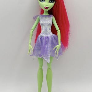 Peut inclure: Une poupée à la peau verte avec de longs cheveux roses vifs et des yeux bleus. La poupée porte une robe blanche et lavande. Les bras et les jambes de la poupée sont articulés, et elle a des oreilles pointues et des crocs. C'est un objet de collection.