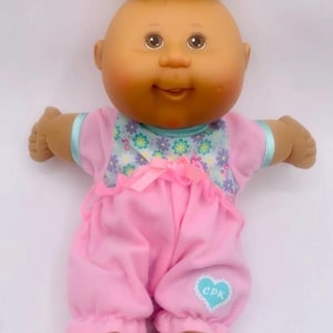 Könnte beinhalten: Eine Vintage-Puppe der Cabbage Patch Kids mit dunkler Haut und schwarzem Haar. Die Puppe trägt ein rosa Outfit mit einem Lätzchen mit Blumenmuster und einem herzförmigen Logo mit der Aufschrift "CPK". Die Arme der Puppe sind ausgestreckt.