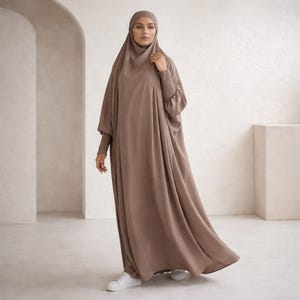 May include: A long, flowing, taupe-colored dress with a matching head covering. The dress features long sleeves with lace detailing at the wrists. The person is wearing white sneakers.