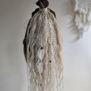 May include: A head of brown and blonde dreadlocks, styled with braids and wavy strands. The dreadlocks are adorned with small beads and feather accents, creating a bohemian aesthetic. The hair is styled in a bun.