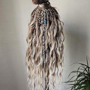 Op de afbeelding: Een pruik met een bruine knot en lange, golvende, blonde en bruine hairextensions. De extensions zijn versierd met kleine blauwe kralen en een zilveren halve maan bedel. Het haar is gestyled op een zwarte standaard.