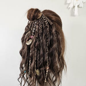 Op de afbeelding: Een bruine pruik gestyled met meerdere vlechten, spiraalaccenten en decoratieve kralen en bedels. De pruik heeft een knot op de kruin en lange, golvende lokken. Het haar is gestyled op een mannequinhoofd.