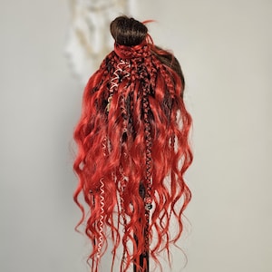 Op de afbeelding: Een felrood haarverlengingsstuk gestyled met losse, vloeiende golven en een knotje bovenop. Het haar bevat ingewikkelde gevlochten secties verweven met dun wit koord, zwarte draad en kleine decoratieve kralen door de gehele lengte.