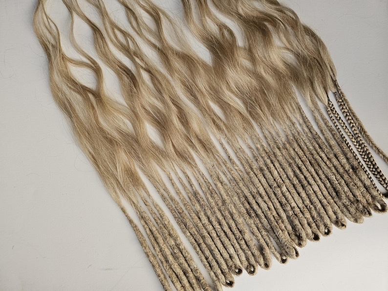 K&ouml;nnte beinhalten: Eine Sammlung blond- und hellbrauner Dreadlocks. Die Dreadlocks gehen von welligem Haar in eng gewickelte, seilartige Str&auml;hnen &uuml;ber. Einige Str&auml;hnen haben einen dunkleren, gemusterten Akzent.