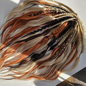 Peut inclure: Ensemble de fausses dreadlocks synthétiques dans diverses nuances de blond, orange, noir et marron. Certaines sont tressées, d'autres lâches, et certaines sont ornées de perles. Les extensions de cheveux sont disposées sur une surface blanche.