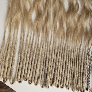 K&ouml;nnte beinhalten: Eine Sammlung von blonden Dreadlocks, mit welligem Haar oben und eng gewickelten, strukturierten Str&auml;hnen unten. Die Dreads sind hellblond, mit einigen Str&auml;hnen, die einen dunkleren, geflochtenen Akzent aufweisen.