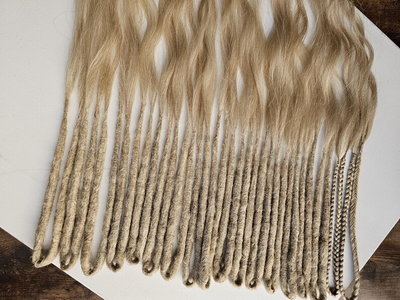 K&ouml;nnte beinhalten: Eine Sammlung von blonden Dreadlocks, mit welligem Haar oben und eng gewickelten, strukturierten Str&auml;hnen unten. Die Dreads sind hellblond, mit einigen Str&auml;hnen, die einen dunkleren, geflochtenen Akzent aufweisen.