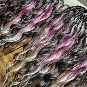 May include: Close-up of several bundles of synthetic dreadlocks. The dreadlocks are a mix of dark gray, pink, and white, with some braids and decorative beads. The wavy texture and color variations create a unique look.