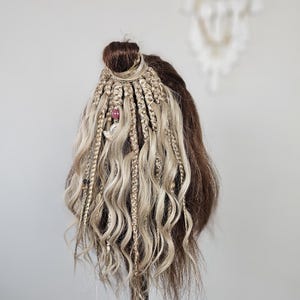 May include: A mannequin head with brown and blonde braided hair, styled in a bun. The braids are decorated with small beads and charms. The hair has wavy, loose strands. The background is a plain, light grey.
