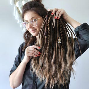 May include: A person holding a collection of brown and blonde dreadlocks. The dreadlocks are decorated with beads and a gold-coloured charm. The person is wearing a plaid shirt and glasses. A dreamcatcher is visible in the background.