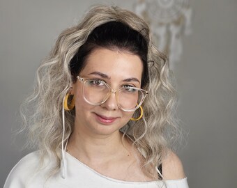 Könnte beinhalten: Eine Person mit einer Brille mit transparentem Rahmen und gelben Creolen. Sie trägt einen weißen Pullover mit dem Wort "sunkissed" in Schreibschrift. Die Person hat welliges blondes Haar und schaut in die Kamera.