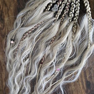 Puede incluir: Un accesorio para el cabello con una base circular y múltiples mechones de cabello sintético ondulado en tono rubio ceniza. Los mechones incluyen varias trenzas, algunas envueltas con hilo oscuro, y están decorados con cuentas y dijes de corazón plateados.