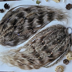 May include: Two sets of brown and blonde dreadlocks with braided and beaded accents. The dreadlocks are arranged on a white surface with decorative elements including pine cones, dried flowers, and a starfish.